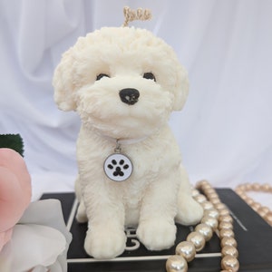 May include: White candle shaped like a sitting poodle dog with a paw print charm around its neck. The candle is sitting on a black book with a pearl necklace.