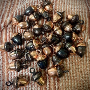 May include: A collection of small, acorn-shaped charms in various metallic finishes. The charms feature a mix of gold, bronze, and dark gray tones, with a loop at the top for attachment. The acorns are scattered on a textured, striped surface.