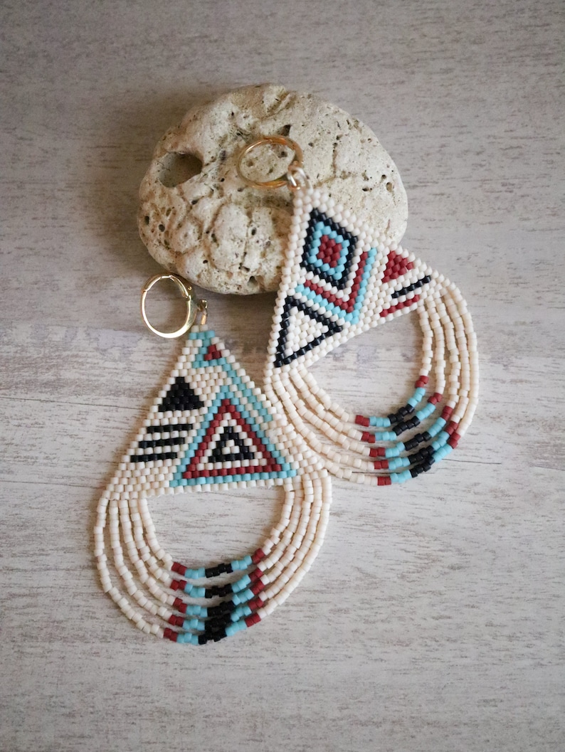 May include: A pair of beaded earrings with a geometric pattern in white, black, red, and blue. The earrings are made of small beads and have a fringe design.