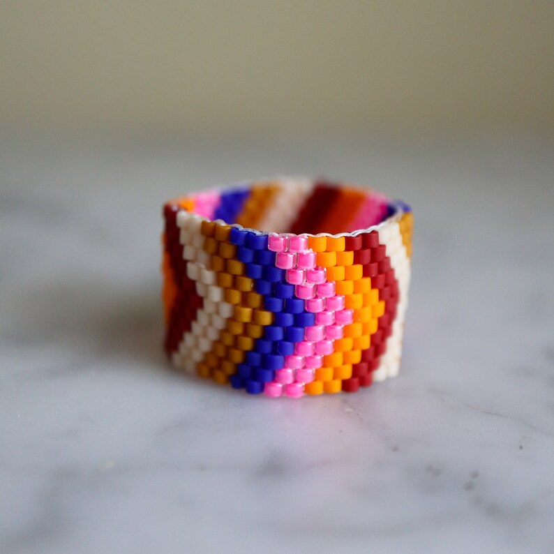 May include: A colorful beaded ring with a chevron pattern in shades of red, orange, yellow, white, pink, and blue.