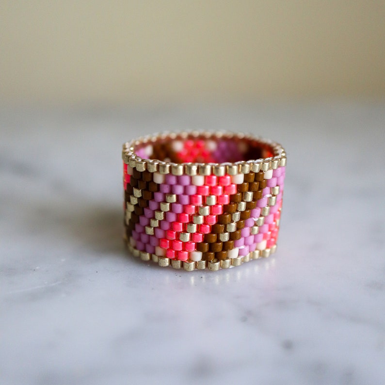 May include: A beaded ring with a geometric pattern in pink, brown, and white colors. The ring is made of small, square beads and has a gold-colored metal band.