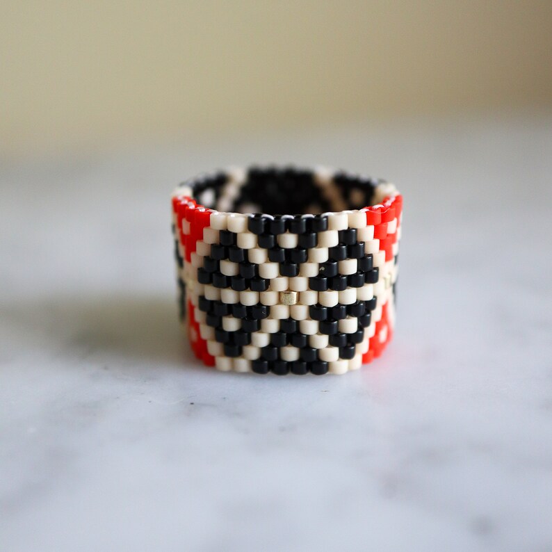 May include: A beaded ring with a geometric pattern in black, white, and red.