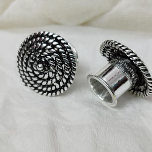 Könnte beinhalten: Zwei silberfarbene Ohrstöpsel. Ein Stöpsel liegt auf der Seite, der andere steht aufrecht. Beide haben ein Spiraldesign auf der Vorderseite und einen seilartigen Rand. Die Stöpsel liegen auf einer weißen Oberfläche.