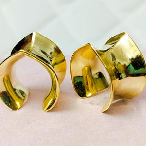 May include: Two gold-colored ear cuffs. The ear cuffs have a curved, open design with a folded, geometric shape at the top. The shiny, metallic finish reflects light, creating highlights and shadows. The earrings are displayed on a pink surface.