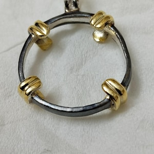 Könnte beinhalten: Ein silberner und goldener Anhänger mit einem runden Rahmen und vier goldenen Akzenten. Der Anhänger ist so konzipiert, dass er eine Münze oder einen anderen kleinen Gegenstand aufnehmen kann.