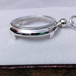 May include: A silver-coloured, oval-shaped pendant with a clear centre, featuring a small loop and a decorative ball. The pendant has a polished, reflective surface and is designed to hold a small item or photo.