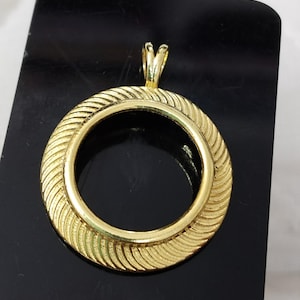 Puede incluir: Un colgante dorado con un diseño circular. El colgante presenta un patrón texturizado y acanalado alrededor del borde exterior y un pequeño lazo en la parte superior para una cadena. El colgante se muestra sobre una superficie negra.