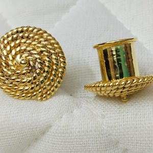 Könnte beinhalten: Zwei goldfarbene Ohrringe im Vintage-Stil. Ein Ohrring ist eine runde Scheibe mit einem seilartigen Design. Der andere hat einen zylindrischen Pfosten und ein seilartiges Design am Sockel. Die Ohrringe liegen auf einer weißen Oberfläche.