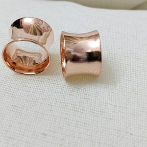 Puede incluir: Dos expansores de oreja de color oro rosa se muestran sobre un fondo blanco. Los expansores tienen un acabado liso y pulido y una forma cilíndrica curvada. Uno está en posición vertical, el otro de lado, revelando el interior hueco.