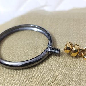 May include: A disassembled metal ring with a screw and a gold-colored clasp. The ring is dark gray, and the clasp has a small ring attached. The objects are on a tan surface.