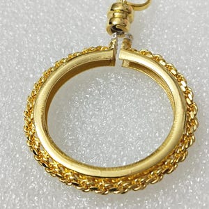 Peut inclure: Un pendentif doré avec un design circulaire. Le pendentif présente une bordure en forme de tresse autour du cercle. Un petit anneau est attaché en haut, et un fermoir magnétique permet d'ouvrir et de fermer le cercle. Le pendentif est sur une surface blanche.