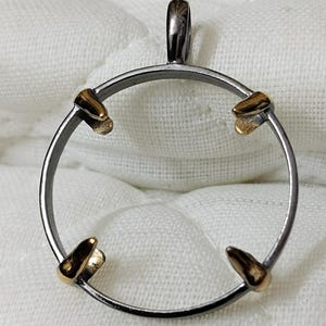 Puede incluir: Un colgante de plata con un marco circular y cuatro acentos dorados. El colgante tiene un bucle en la parte superior para sujetar una cadena.