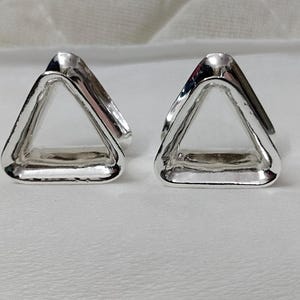 May include: Two silver-colored, triangular-shaped objects. Each object has a hollow center and a smooth, reflective surface. The objects are placed side by side on a white surface, with soft lighting creating highlights and shadows.