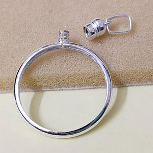 May include: A silver-colored, circular pendant base with a small, detached clasp. The pendant has a loop at the top for attaching a chain. The clasp is cylindrical with a small rectangular loop. The items are on a beige surface.
