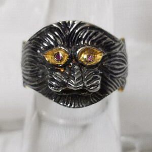 Può includere: Un anello d'argento con un designo dettagliato di un viso di leone. Gli occhi del leone sono incastonati con pietre preziose rosa e accenti dorati.