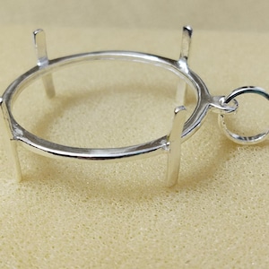 May include: A silver metal pendant setting with a circular design. It has four vertical prongs and a small loop for a chain. The setting is designed to hold a round gemstone or other decorative element.
