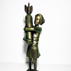 May include: A bronze-colored sculpture of a young person embracing a bomb. The figure is wearing a dress and shoes, and the sculpture is mounted on a square base. The artwork has a distressed finish.