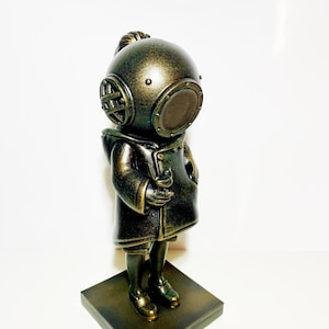 May include: A bronze-colored figurine of a person wearing a diving helmet and a long coat. The figurine is standing on a square base.
