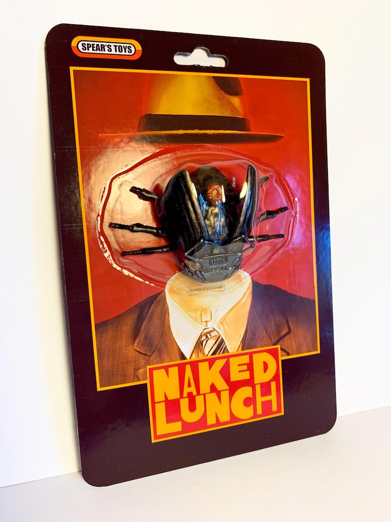 Puede incluir: Figura de acci&oacute;n de un escarabajo con sombrero y traje, empaquetada en una tarjeta retro. La tarjeta presenta el texto "NAKED LUNCH" en naranja y el logotipo "SPEAR'S TOYS". El embalaje es de color granate oscuro.