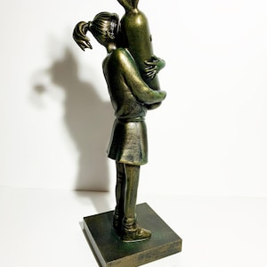 May include: A bronze-colored sculpture of a young person hugging a bomb. The figure is wearing a skirt and has their hair in a ponytail. The sculpture is mounted on a square base.