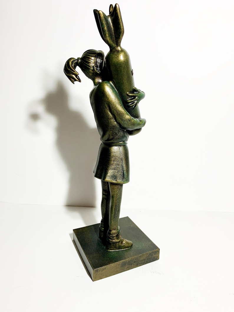 May include: A bronze-colored sculpture of a young person hugging a bomb. The figure is wearing a skirt and has their hair in a ponytail. The sculpture is mounted on a square base.