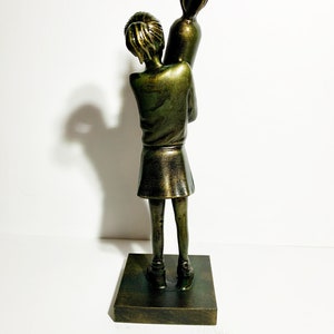 May include: A bronze-colored sculpture of a young person holding a large, stylized object resembling a rocket. The figure is wearing a skirt and top, standing on a square base. The sculpture has a distressed finish.