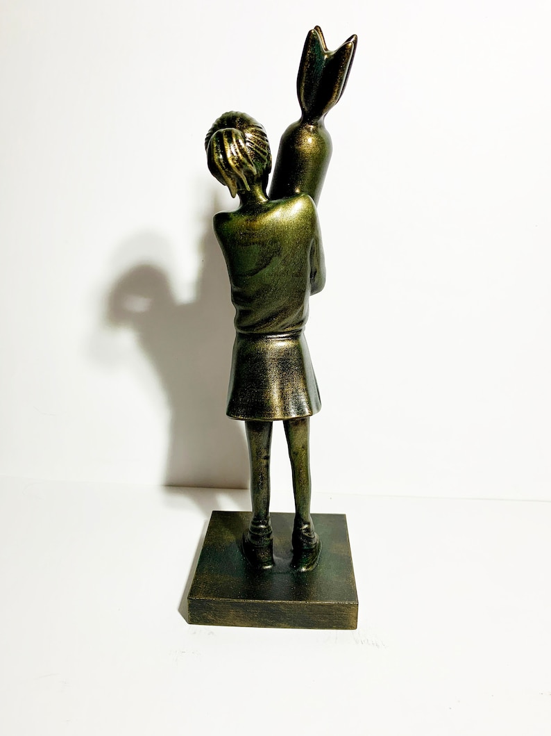 May include: A bronze-colored sculpture of a young person holding a large, stylized object resembling a rocket. The figure is wearing a skirt and top, standing on a square base. The sculpture has a distressed finish.
