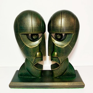 Könnte beinhalten: Ein Paar abstrakter Gesichtsskulpturen in Grün und Gold. Die Skulpturen stehen nebeneinander auf einem rechteckigen Sockel. Die Gesichter haben große, stilisierte Augen und offene Münder. Die Farbe ist eine Mischung aus Grün und Gold.