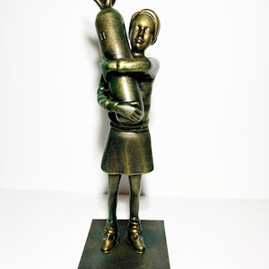 May include: A bronze-colored sculpture of a child hugging a bomb. The figure is standing on a square base. The child is wearing a short skirt and shoes. The sculpture has a metallic finish and is set against a white background.