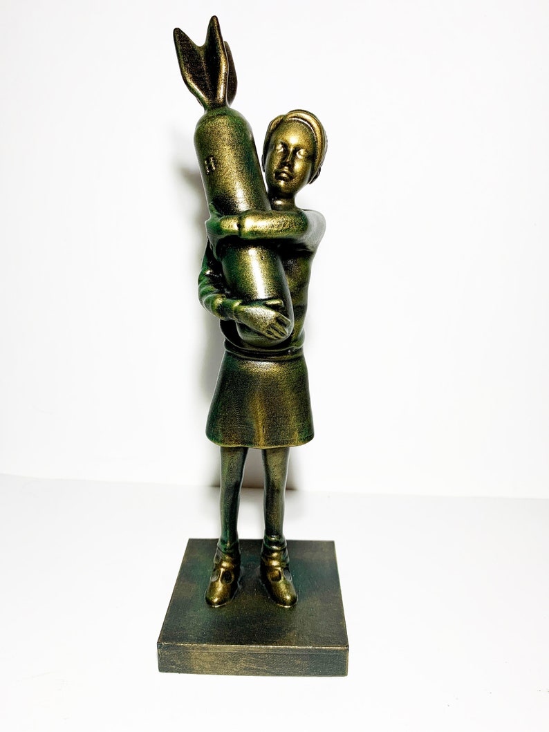 May include: A bronze-colored sculpture of a child hugging a bomb. The figure is standing on a square base. The child is wearing a short skirt and shoes. The sculpture has a metallic finish and is set against a white background.