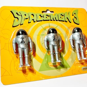 May include: A set of three silver Spacemen 3 action figures on a yellow and green card with the text "Spacemen 3" in a retro font. The figures are wearing silver spacesuits with black helmets and a triangle with a laser beam symbol on their chests. The card also has the text "Speak's Toys" in the bottom right corner.