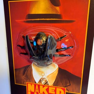 Puede incluir: Un paquete de juguetes con una criatura negra con forma de ara&ntilde;a con ojos azules, un sombrero amarillo y el texto "NAKED LUNCH". El paquete tiene el logotipo "SPEAR'S TOYS".