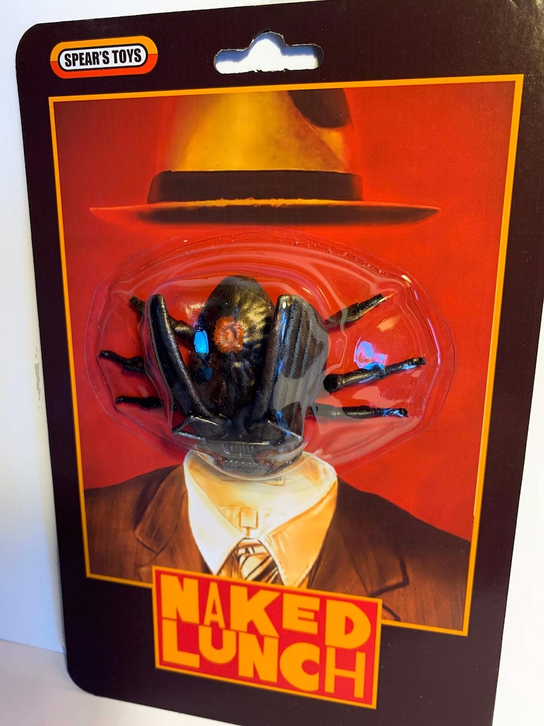 Puede incluir: Un paquete de juguetes con una criatura negra con forma de ara&ntilde;a con ojos azules, un sombrero amarillo y el texto "NAKED LUNCH". El paquete tiene el logotipo "SPEAR'S TOYS".