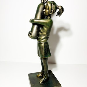 May include: A bronze-colored sculpture of a young person hugging a large rabbit. The figure is wearing a skirt and shoes, and the sculpture is mounted on a dark wooden base. The rabbit has long ears.