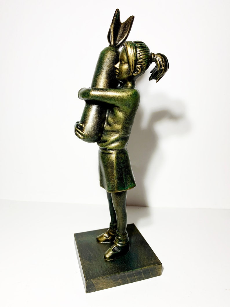May include: A bronze-colored sculpture of a young person hugging a large rabbit. The figure is wearing a skirt and shoes, and the sculpture is mounted on a dark wooden base. The rabbit has long ears.