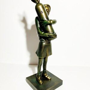 May include: A bronze-colored sculpture of a young person holding a bomb. The figure is wearing a dress and boots, standing on a square base. The bomb has a fin and the letter "H" on it. The sculpture has a metallic finish.