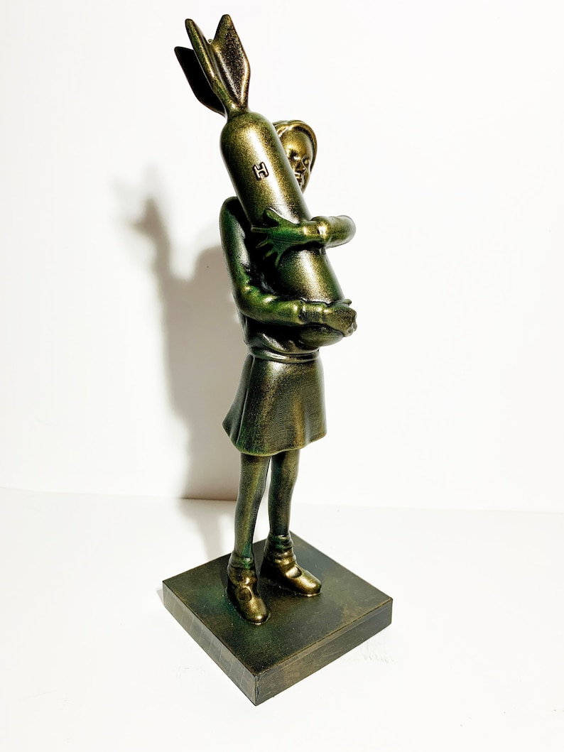 May include: A bronze-colored sculpture of a young person holding a bomb. The figure is wearing a dress and boots, standing on a square base. The bomb has a fin and the letter "H" on it. The sculpture has a metallic finish.