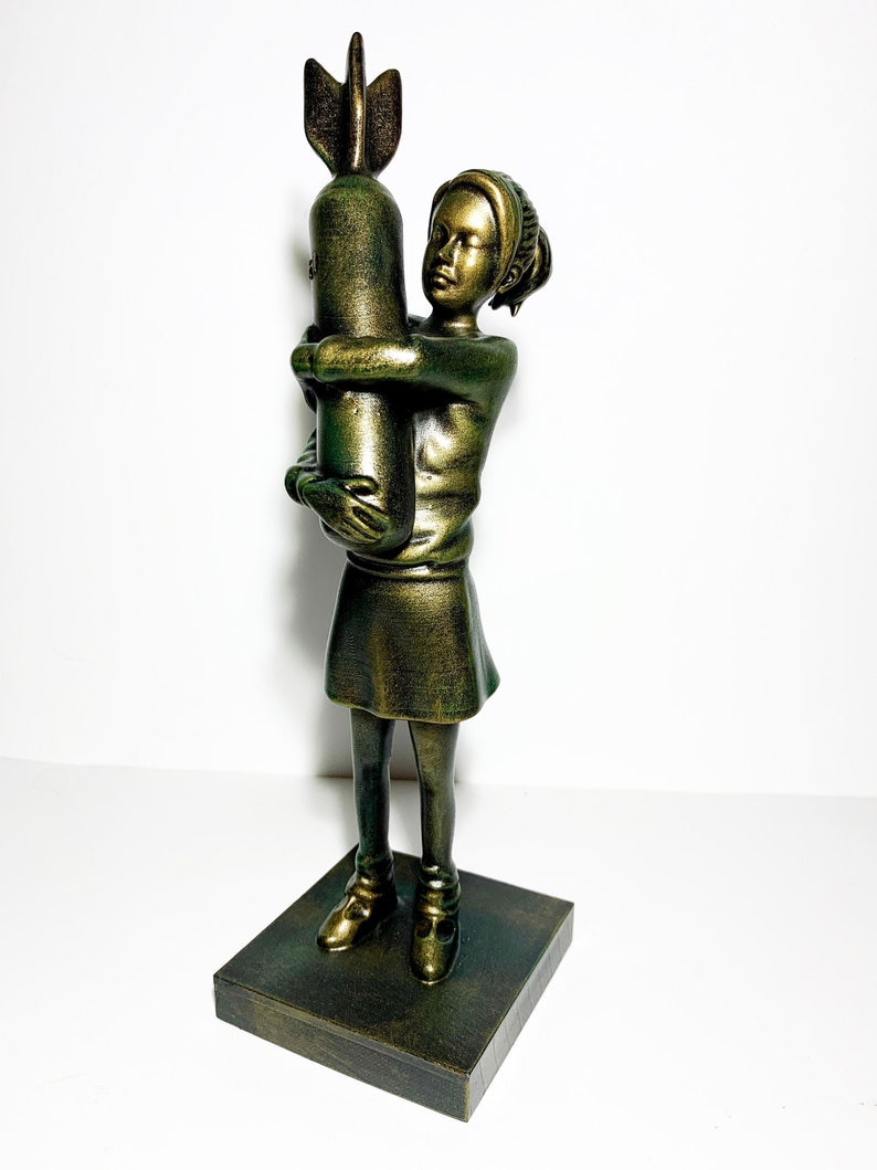 May include: A bronze-colored sculpture of a young person embracing a bomb. The figure is wearing a dress and shoes, and the sculpture is mounted on a square base. The artwork has a distressed finish.