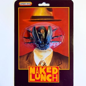 Puede incluir: Paquete de juguete con una cabeza de insecto negro con sombrero fedora y traje. El paquete tiene el texto "NAKED LUNCH" en amarillo y rojo. La marca "SPEAR'S TOYS" est&aacute; en la parte superior del paquete.