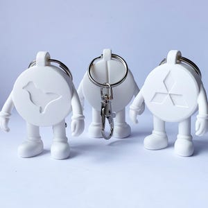 Könnte beinhalten: Drei weiße Schlüsselanhänger in Form von Cartoonfiguren. Jede Figur hat einen runden Körper mit einem anderen Design: ein Vogel, ein leerer Kreis und ein Drei-Diamanten-Logo. Die Schlüsselanhänger haben silberne Schlüsselringe und Verschlüsse.