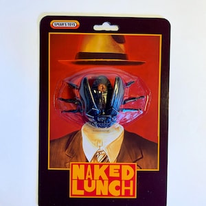 Puede incluir: Un paquete de figura de acci&oacute;n de estilo vintage con una cabeza de insecto negra y un sombrero fedora sobre un traje. El texto "NAKED LUNCH" est&aacute; en naranja. La marca "SPEAR'S TOYS" est&aacute; en la parte superior.