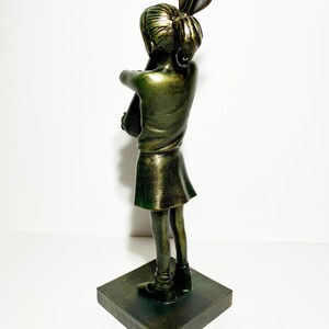 May include: A bronze-colored sculpture of a girl with her arms crossed, standing on a square base. The girl's hair is styled in a high bun with a decorative element. The sculpture has a metallic finish and is set against a white background.