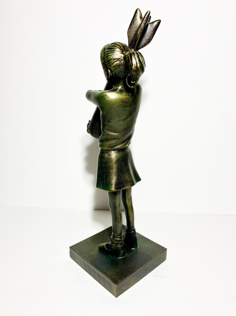 May include: A bronze-colored sculpture of a girl with her arms crossed, standing on a square base. The girl's hair is styled in a high bun with a decorative element. The sculpture has a metallic finish and is set against a white background.