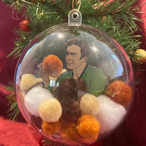 May include: A clear glass Christmas ornament with a cartoon image of a man inside. The ornament is filled with white, brown, and orange pom-poms.
