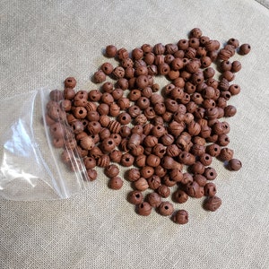 Op de afbeelding: Een grote hoeveelheid bruine klei kralen met een getextureerd oppervlak. De kralen zitten in een doorzichtige plastic zak op een lichtbruin stoffen oppervlak.