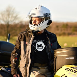 Op de afbeelding: Een persoon met een witte helm en een zwart T-shirt met een motorfietsafbeelding en de tekst "Overlander" zit op een gele Suzuki motorfiets. De motorfiets heeft zwarte zadeltassen en een zwarte topkoffer.