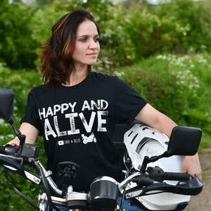 May include: A woman wearing a black t-shirt with white text that reads "Happy and Alive" and a motorcycle helmet sits on a motorcycle. The t-shirt also has a small graphic of a motorcycle with the text "Lavi & Ollie".