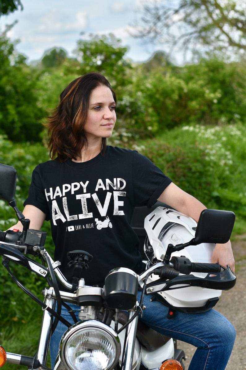 May include: A woman wearing a black t-shirt with white text that reads "Happy and Alive" and a motorcycle helmet sits on a motorcycle. The t-shirt also has a small graphic of a motorcycle with the text "Lavi & Ollie".