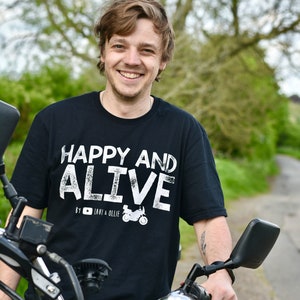May include: A man wearing a black t-shirt with white text that reads "Happy and Alive" by "Lavi & Ollie" is sitting on a black motorcycle. He is smiling and looking at the camera.