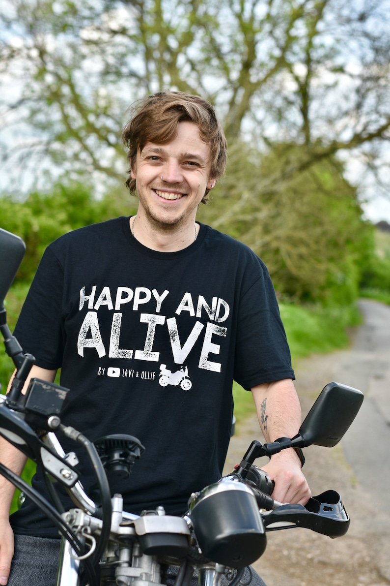 May include: A man wearing a black t-shirt with white text that reads "Happy and Alive" by "Lavi & Ollie" is sitting on a black motorcycle. He is smiling and looking at the camera.